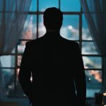 Mies seisoo pimeässä huoneessa vasten ikkunaa, jonka takaa heijastuvat öiset kaupungin valot. Miestä ei tunnista, hän on vain tumma varjo kuvassa.